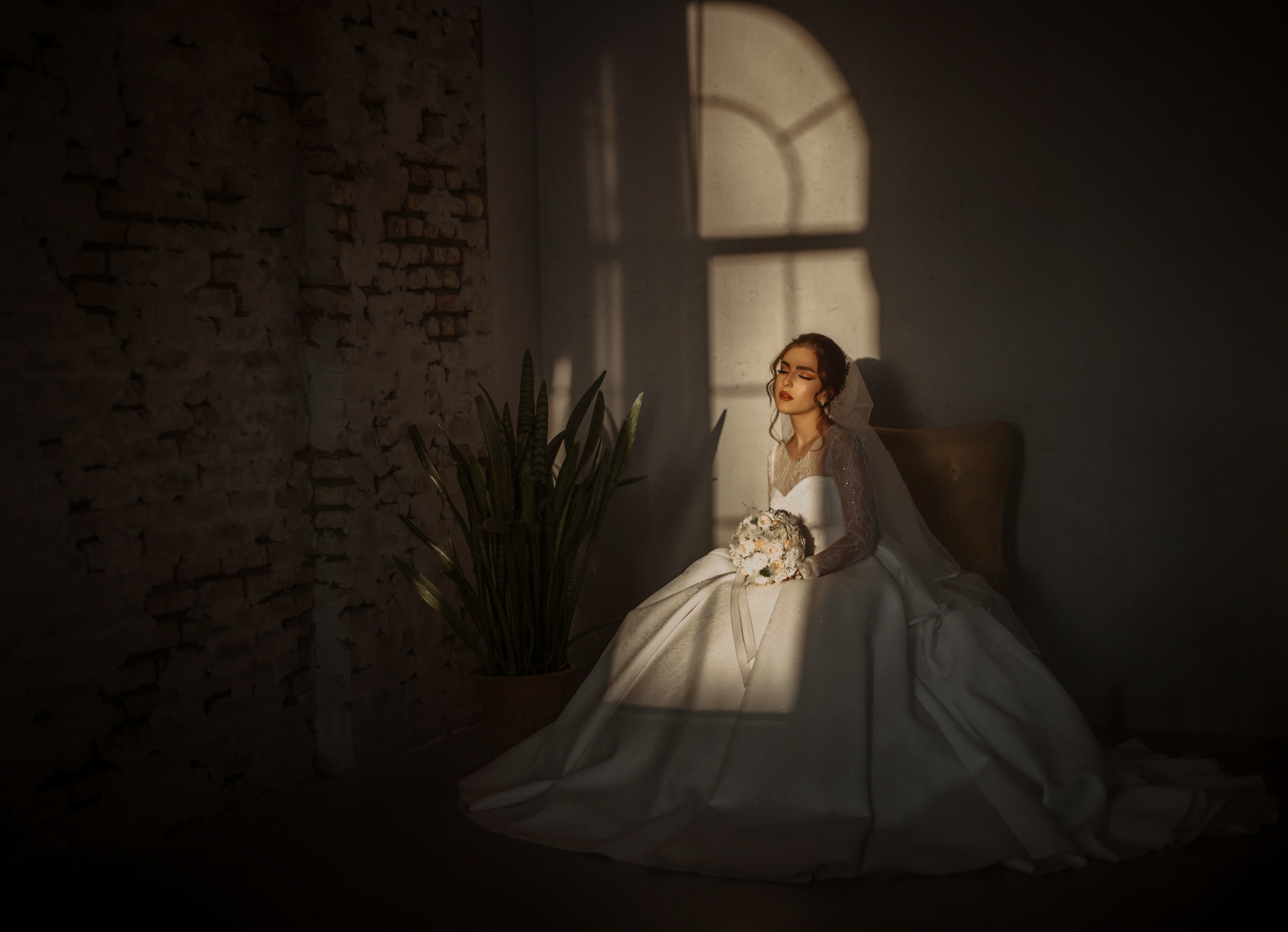 A bride sits in soft window light in a lace-sleeved gown and veil, holding a pale bouquet